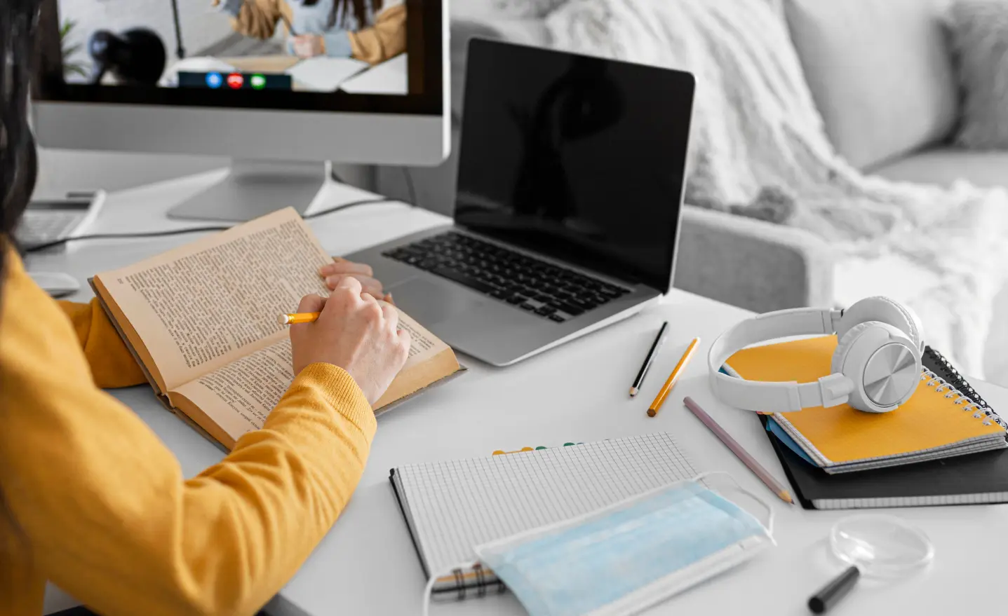 Fundo com pessoa em mesa de trabalho com um livro na mão em frente a um computador.
